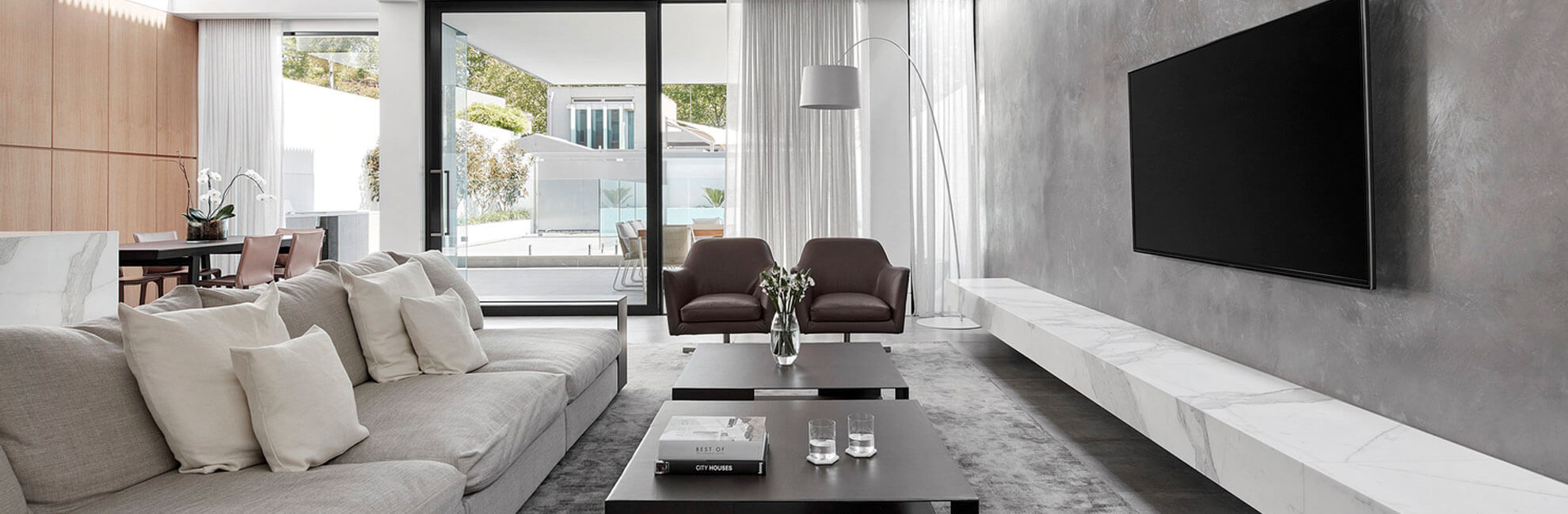 Wide banner of a modern living room with grey sofa, low marble-topped media wall and glass doors to a pool terrace.
